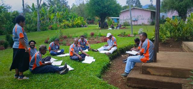 Facilitators in programme shirts sit in small groups on a lawn, working through materials.
