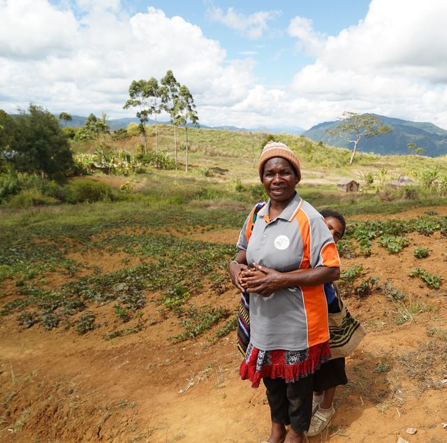 Margaret Lord, a Community Curriculum facilitator, stands in a Highlands garden with a child at her back, mountains behind her.